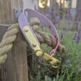 A pastel yellow and lavender dog collar with brass fittings hangs on a thick rope tied to a wooden post, surrounded by blooming lavender plants at sunset.