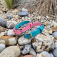 Turquoise and pink custom two-colour BioThane dog collars with contrasting buckles, displayed on natural pebbles and gravel.