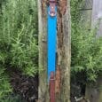 The Driftwood Tapered Collar is shown upright on a weathered wooden post in a garden, surrounded by greenery and pebbles. It features blue and brown leather with brass fittings.