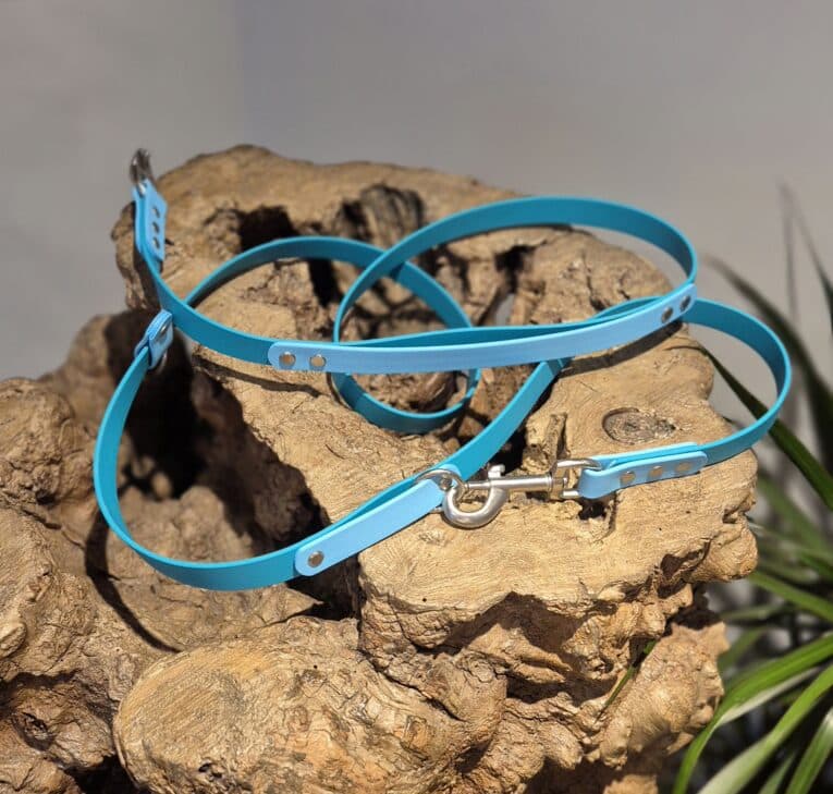 The Custom 2m Multi-way Double-ended Lead, featuring bright blue material and silver metal fittings, is coiled on textured driftwood with green leaves visible in the background.