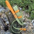 A Custom Two Colour 1.2m Lead dog collar and lead set in green and orange rests on rocks and wood, surrounded by lush plants in a garden setting.