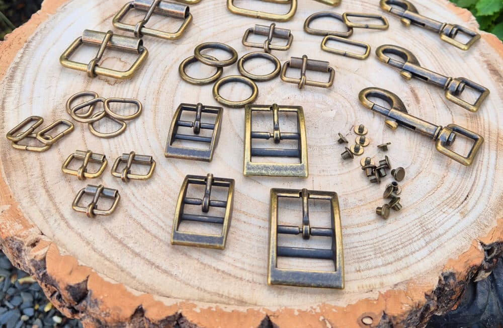 Various brass-coloured metal buckles, rings, and clasps arranged neatly on a tree stump slice—perfect hardware for sustainable dog collars UK—set outdoors with greenery and stones in the background.