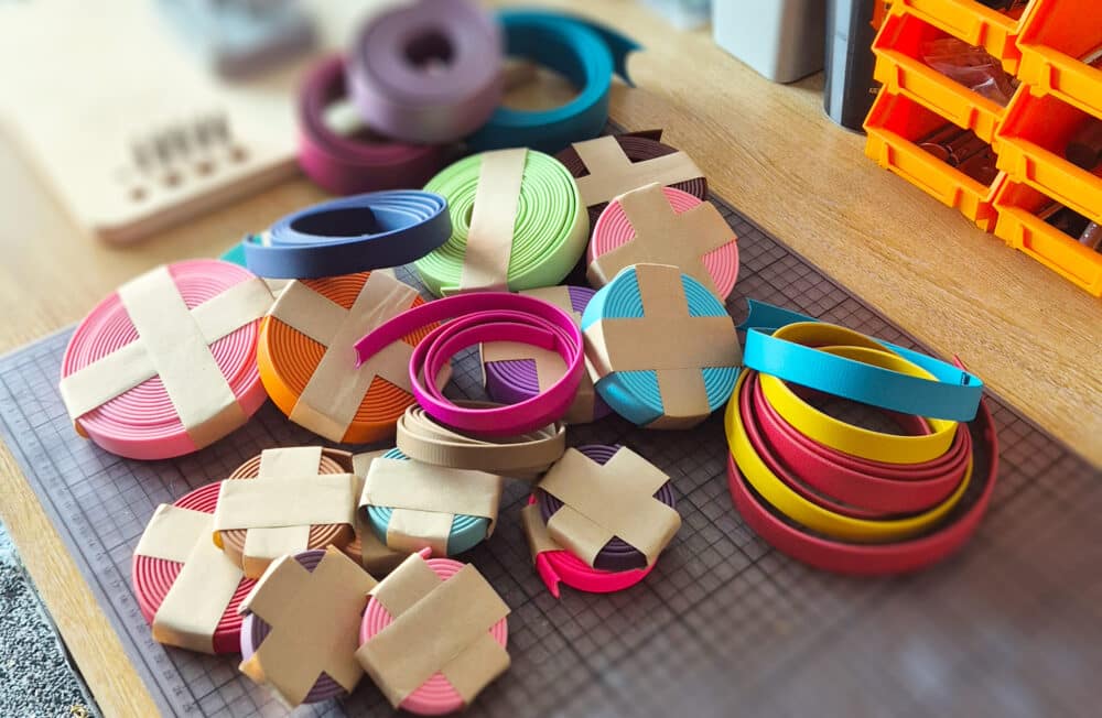 Colourful rolls of paper strips and bands, some wrapped with brown paper, are neatly arranged on a wooden desk alongside craft supplies—an inspiring scene for makers of sustainable dog collars UK. Storage boxes sit in the background.