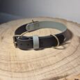 A black and grey leather collar with gold fittings rests on a round wooden surface with visible tree rings.