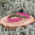 Two pink dog collars with gold buckles are displayed on a round wooden slab, surrounded by silvery-green foliage and lavender stems in an outdoor setting.