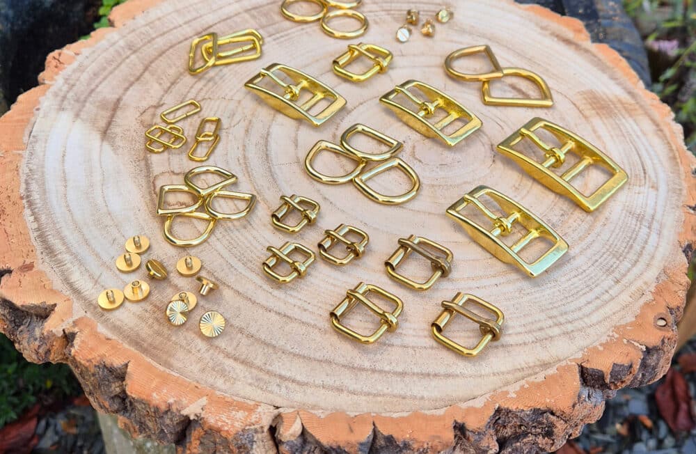 A variety of shiny brass buckles, rings, and rivets for sustainable dog collars UK are neatly arranged on a large tree stump with visible rings, outdoors near greenery and gravel.