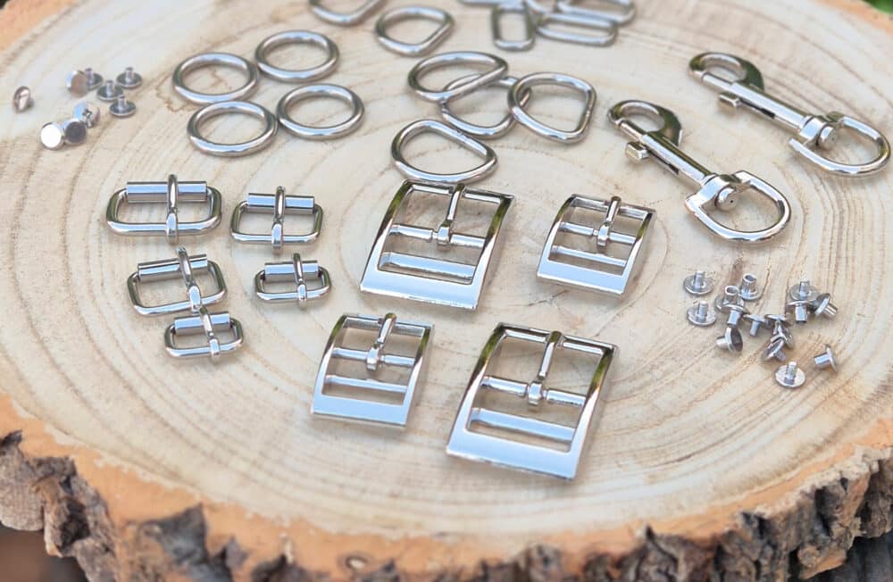 Assorted metal buckles, rings, clasps, and rivets—often used in sustainable dog collars UK—are neatly arranged on a round wooden slab outdoors, with greenery blurred in the background.