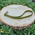 A Custom Single Colour Traffic Handle in olive green with a metal clasp rests on a round wooden tree stump, surrounded by green leafy plants.