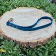 The Custom Single Colour Traffic Handle lead, featuring a metal clasp, is laid in a loop on a round wooden tree slice and surrounded by green leafy plants.