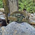 A Custom Quick-Snap Bungee Collar Strap in tan, featuring metal rivets and a black circular ring, lies on a grey rock outdoors among plants, other rocks, and a weathered wooden post.