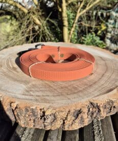 The Waterproof Hexa Long-Line / Training Lead, a coiled orange lead with a honeycomb pattern and customisable traffic handle, rests on a large circular wooden slab outdoors amid lush trees and greenery.