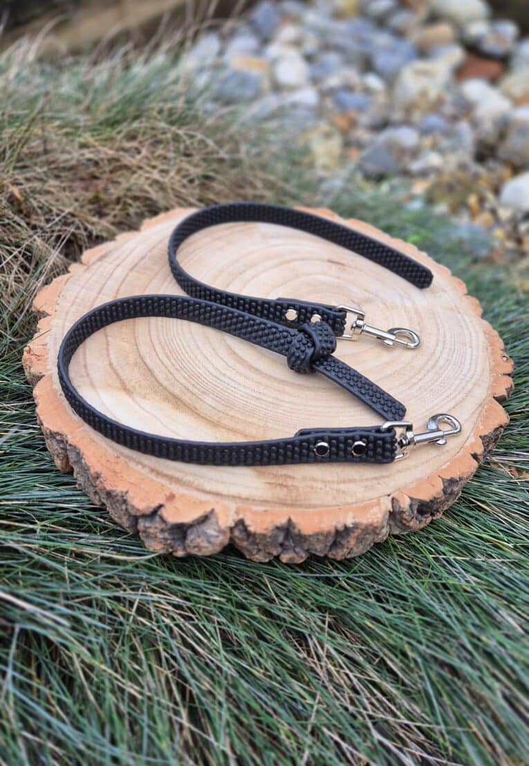 A black, textured Super-grip Biothane Training Tab/Obedience Lead - 51cm/20in with metal clasps is coiled on a round wooden slab placed on green grass, surrounded by rocks in the background.