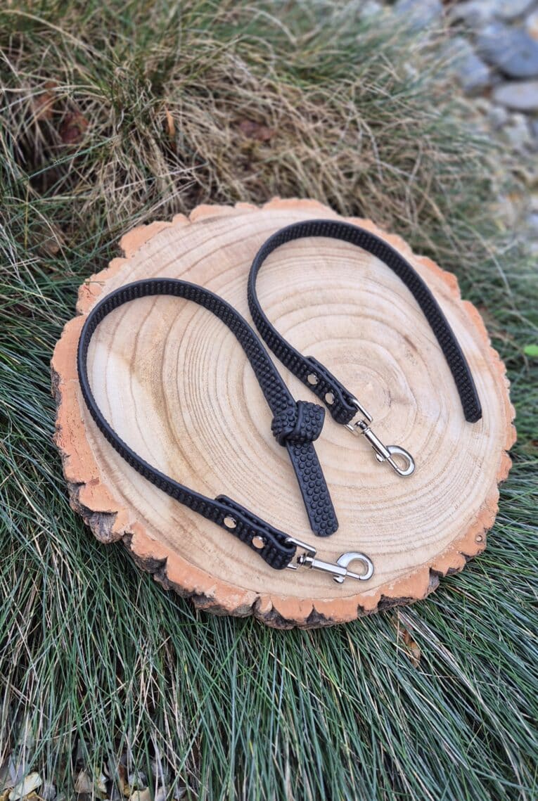 Two Super-grip Biothane Training Tabs/Obedience Leads (51cm/20in) with metal clasps rest on a round wooden slice in green grass—customisable, durable tabs designed for added control and style.