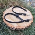 Two black, textured Super-grip Biothane Training Tab/Obedience Leads (51cm/20in), each with a metal clip, are coiled on a round tree stump outdoors among grass, rocks, and green plants.