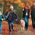 Two children run ahead on a leaf-covered path, smiling, while two adults walk behind them holding hands and walking a dog with a sturdy Biothane collar. The scene is set in an autumn forest with colourful foliage.