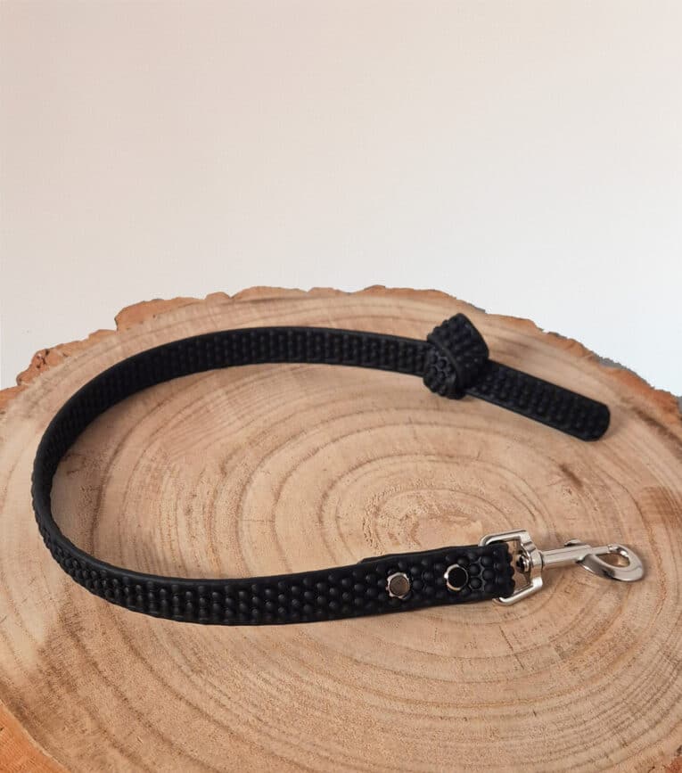 The Super-grip Biothane Training Tab / Obedience Lead 51cm, a short black textured dog lead with a metal clasp and customisable traffic handle, is coiled on a round light wooden surface against a plain white background.