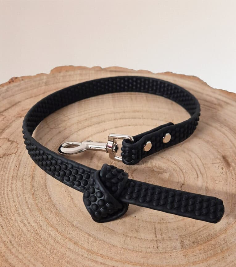 A Super-grip Biothane Training Tab / Obedience Lead 51cm with a black textured finish and silver metal clasp is neatly coiled on a round, light-coloured wooden surface.