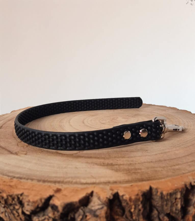 A black textured Super-grip Biothane Training Tab / Obedience Lead 51cm with metal buckle and holes rests on a round wooden surface, alongside a customisable traffic handle lead, set against a plain light background.