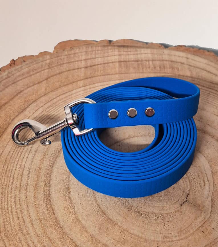 A coiled bright blue Waterproof Hexa Long-Line / Training Lead with a metal clasp and three silver rivets, shown on a round wooden surface with visible tree rings.