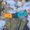 A yellow and teal Quick-Snap Bungee Collar with a black buckle rests on a weathered tree stump outdoors, surrounded by rocks, green plants, and patches of snow.