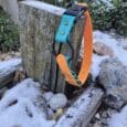 The Quick-Snap Bungee Collar, featuring a teal and orange strap, hangs on a weathered wooden post outdoors amid rocks, greenery, and a light layer of snow.