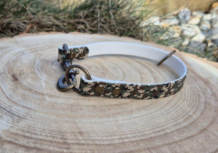 The Custom Snap Tag - Slip Collar, featuring a quick-snap design and brass fittings in a camouflage pattern, is shown on a tree stump with grass and stones in the background.