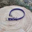 A Custom Snap Tag/Slip Collar in purple with silver hardware rests on a round wooden surface outdoors, surrounded by grass and stones.