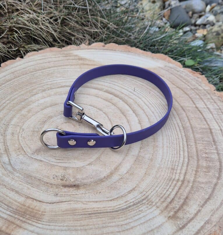 A Custom Snap Tag/Slip Collar in purple with silver hardware rests on a round wooden surface outdoors, surrounded by grass and stones.