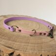 A two-tone dog collar in pastel purple and pink with brass fittings is displayed on a round, wooden surface with visible tree rings.