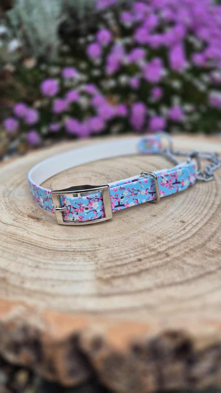 Handmade BioThane dog collar with bright blue floral pattern and silver buckle, displayed on wooden surface with purple flowers blurred behind.