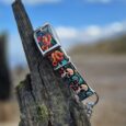 Handmade BioThane dog collar with vibrant floral skull design displayed on weathered wooden post against blue sky.