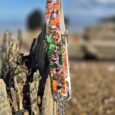 Colourful handmade BioThane dog lead with vibrant floral pattern attached to weathered wooden post on beach.