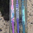 Three waterproof printed dog collars in pink, purple, and teal displayed on weathered wood, showcasing vibrant designs and quick-snap hardware.