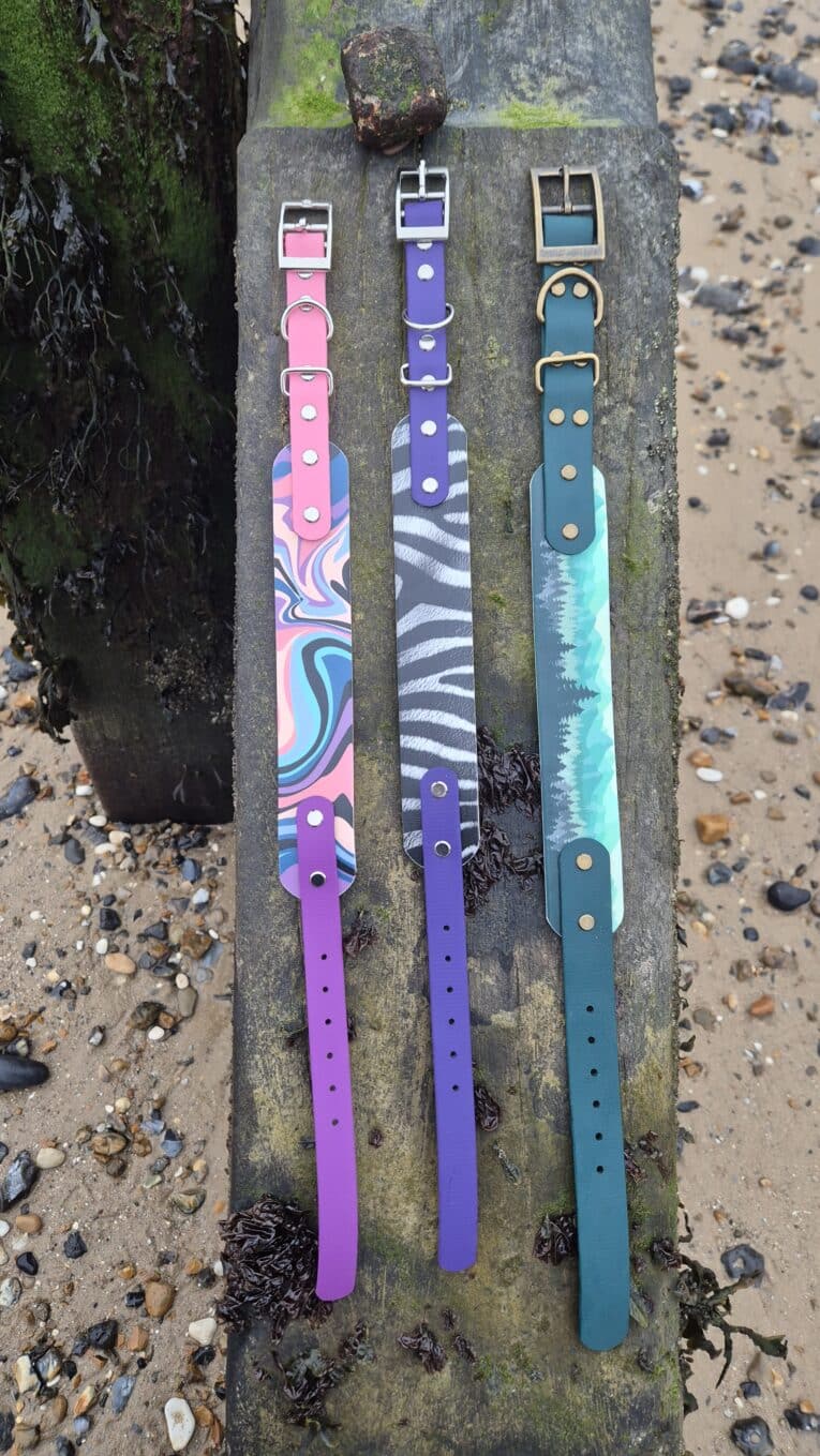 Three waterproof printed dog collars in pink, purple, and teal displayed on weathered wood, showcasing vibrant designs and quick-snap hardware.