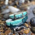 Handmade BioThane dog collar in turquoise with forest tree design, displayed on coastal pebbles and rocks.