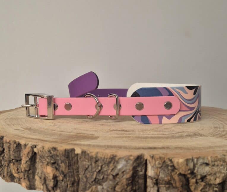 Pink waterproof BioThane dog collar with quick-snap hardware displayed alongside purple and patterned printed hound collar options on wooden surface.