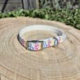 Colourful handmade BioThane dog collar with pink, blue and yellow pattern, displayed on wooden surface with garden background.