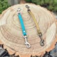 Two quick-snap customisable dog collars in turquoise and tan Biothane displayed on a wooden cross-section, featuring metal hardware and adjustable fittings.