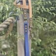 Handmade BioThane dog lead in blue with tan handle, displayed on wooden post surrounded by garden foliage.