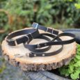 Three black handmade BioThane dog collars with metal hardware displayed on a wooden log outdoors.