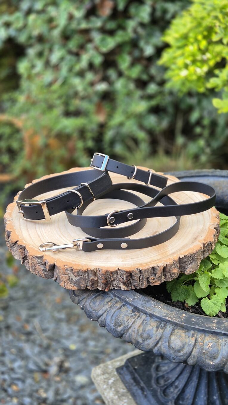 Three black handmade BioThane dog collars with metal hardware displayed on a wooden log outdoors.