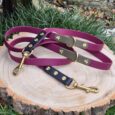 Three handmade BioThane dog leads in purple, charcoal, and navy with gold hardware, displayed on wood slice.