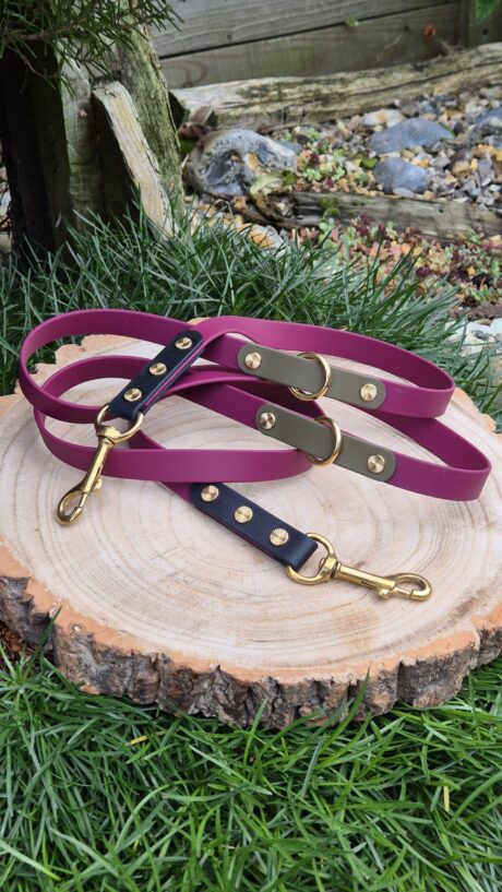 Three handmade BioThane dog leads in purple, charcoal, and navy with gold hardware, displayed on wood slice.