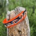 Vibrant orange handmade BioThane dog collar with dragon design wrapped around weathered wooden post outdoors.