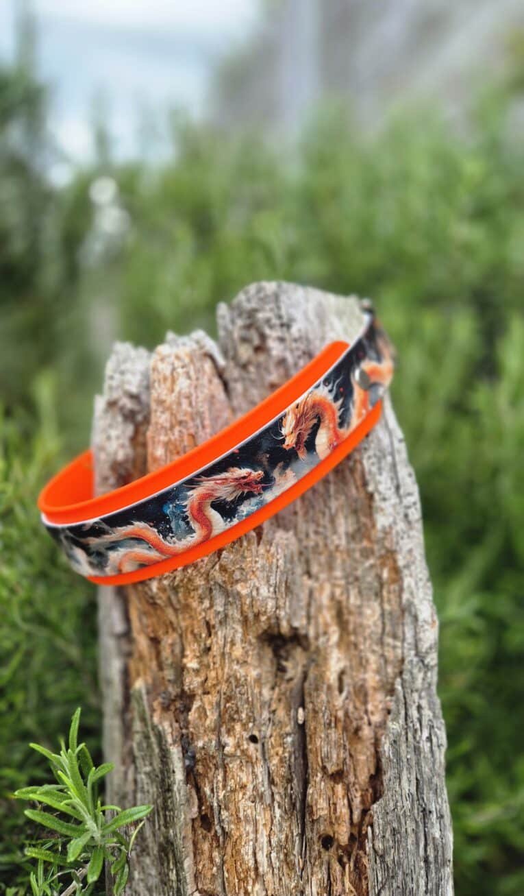Vibrant orange handmade BioThane dog collar with dragon design wrapped around weathered wooden post outdoors.