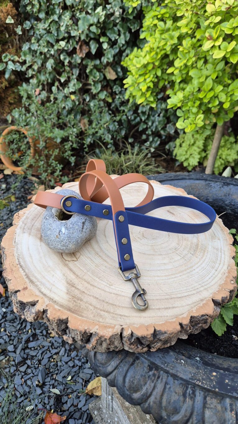 Two handmade BioThane dog leads displayed on a wood slice: navy blue with brass hardware and tan brown, set in a garden.