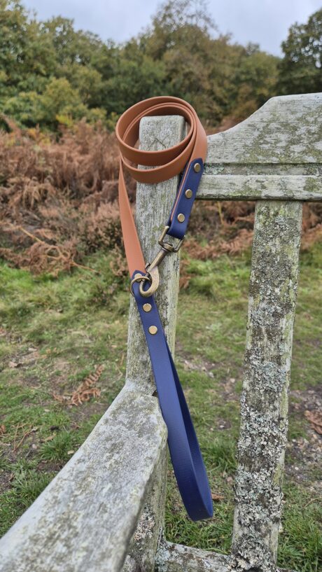 Tan BioThane dog lead with navy handle and brass fittings, draped over weathered wooden fence.