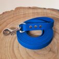 Bright blue handmade BioThane dog longline coiled on natural wood, featuring silver hardware and studded collar detail.
