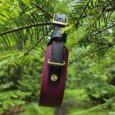 Handmade BioThane dog collar with burgundy and black design, brass hardware, suspended among green foliage.