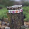 Two handmade BioThane dog collars stacked on weathered wood: camouflage design with studs and white printed collar with colourful heart motifs.
